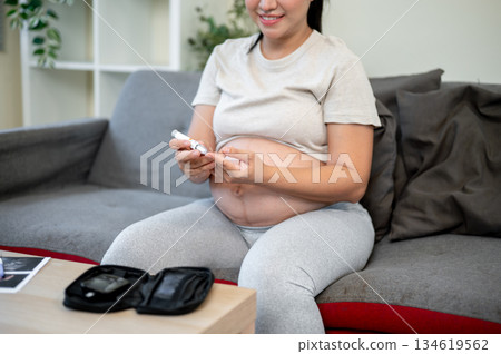 Pregnant asian woman with bare belly drawing a drop of finger blood for glucose testing kit on sofa. 134619562