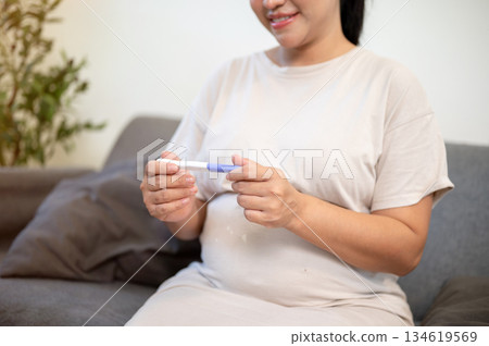 Pregnant asian woman holding and looking at a pregnancy test kit while sitting on sofa in the house. Pregnant asian woman holding and looking at a pregnancy test kit while sitting on sofa in the house. 134619569