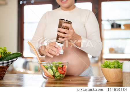 Close up of pregnant asian woman grinding salt into glass bowl making a vegetables salad in kitchen. Close up of pregnant asian woman grinding salt into glass bowl making a vegetables salad in kitchen. 134619584
