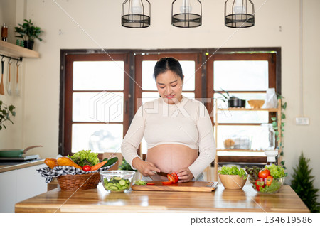 Pregnant asian woman is holding knife cutting a tomato on chopping board making salad in the kitchen Pregnant asian woman is holding knife cutting a tomato on chopping board making salad in the kitchen 134619586