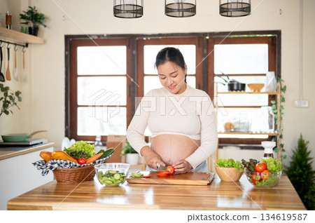 Pregnant asian woman is cutting a tomato with knife on chopping board making salad in the kitchen. Pregnant asian woman is cutting a tomato with knife on chopping board making salad in the kitchen. 134619587