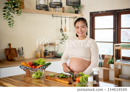 Pregnant asian woman smiling as she cutting cucumbers with knife on chopping board in the kitchen. Pregnant asian woman smiling as she cutting cucumbers with knife on chopping board in the kitchen. 134619589