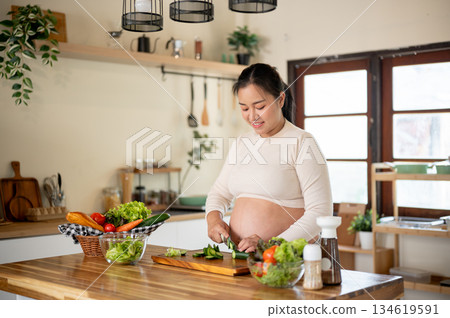 Pregnant asian woman holding knife chopping cucumbers on cutting board preparing food in the kitchen Pregnant asian woman holding knife chopping cucumbers on cutting board preparing food in the kitchen 134619591