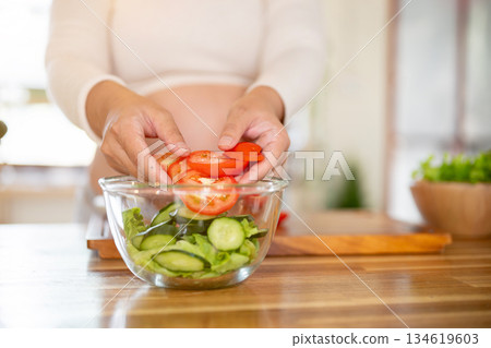 Close up of pregnant woman putting tomato slices in glass bowl making a vegetables salad in kitchen. Close up of pregnant woman putting tomato slices in glass bowl making a vegetables salad in kitchen. 134619603