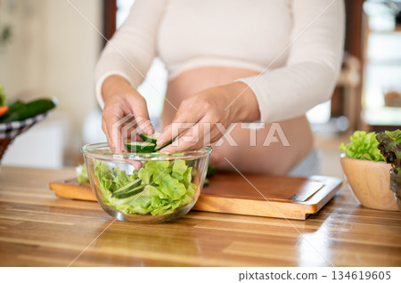 Close up of pregnant woman putting cucumber slices into glass bowl making vegetables salad in kitchen. Close up of pregnant woman putting cucumber slices into glass bowl making vegetables salad in kitchen. 134619605
