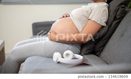 Close up of pregnant asian woman holding bare belly in both hands sitting leaning on sofa in house. Close up of pregnant asian woman holding bare belly in both hands sitting leaning on sofa in house. 134619609