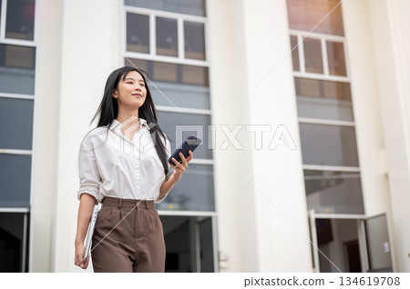 Asian woman in shirt holding phone looking ahead while walking out of university or office building. Asian woman in shirt holding phone looking ahead while walking out of university or office building. 134619708