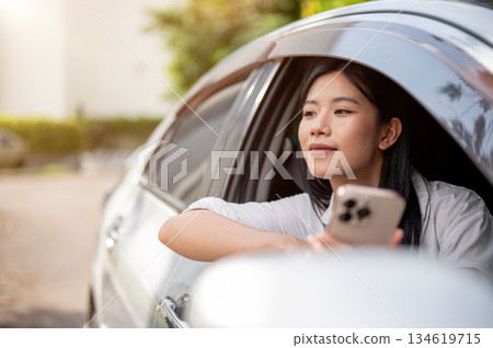 Asian woman in white shirt looking away from phone inside a car parked at parking lot or roadside. Asian woman in white shirt looking away from phone inside a car parked at parking lot or roadside. 134619715