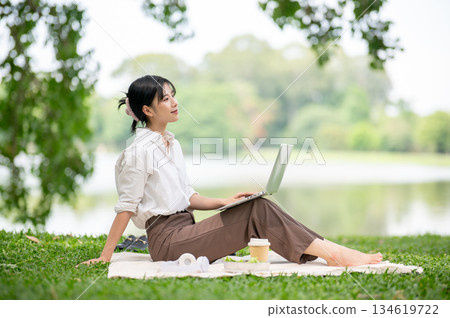 Asian woman in white shirt looking away from her laptop while sitting knee up on picnic mat laying on grass. Asian woman in white shirt looking away from her laptop while sitting knee up on picnic mat laying on grass. 134619722