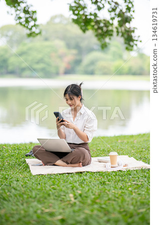 Asian woman in white shirt looking at phone holding in both hands sitting on picnic mat lay on grass 134619731