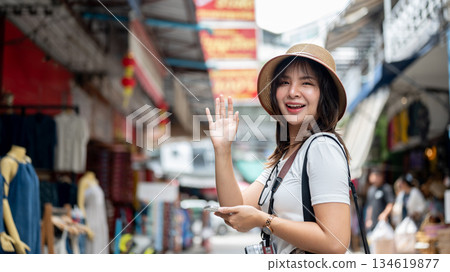 Asian woman wearing hat smiling while waving her hand standing on sidewalk in the local old market. Asian woman wearing hat smiling while waving her hand standing on sidewalk in the local old market. 134619877
