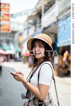 Asian woman wearing hat smiling while holding her phone standing on sidewalk in the local old market 134619878