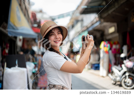 Asian woman wearing hat smiling while holding a camera taking photos in the local old market. Asian woman wearing hat smiling while holding a camera taking photos in the local old market. 134619889
