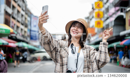 Asian tourist woman holding phone pointing finger in video call standing by the road of local market Asian tourist woman holding phone pointing finger in video call standing by the road of local market 134620008