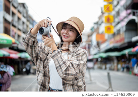 Asian tourist woman wearing hat holding camera taking photos while standing by the road of local market. Asian tourist woman wearing hat holding camera taking photos while standing by the road of local market. 134620009