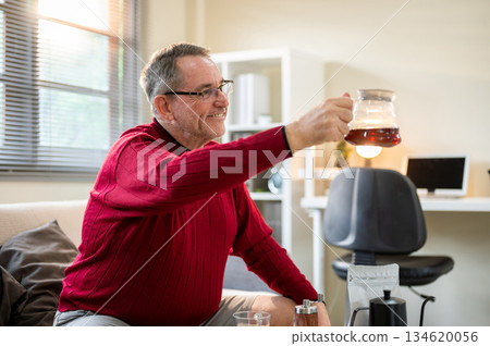 Old white caucasian man wearing glasses showing his coffee in the dripper on sofa in a living room. Old white caucasian man wearing glasses showing his coffee in the dripper on sofa in a living room. 134620056