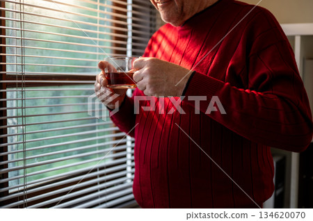 Close up of old white caucasian man holding coffee in glass cup and standing near the window blinds. 134620070