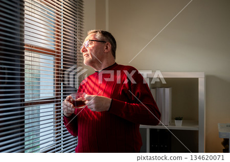 Old white caucasian man wearing glasses holding his coffee cup and looking out through window blinds 134620071