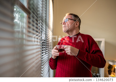 Old white caucasian man wearing glasses holding his coffee cup looking ahead through window blinds. 134620073