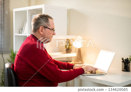 Glasses old white caucasian man typing and looking at laptop with lamp aside on table in living room 134620085