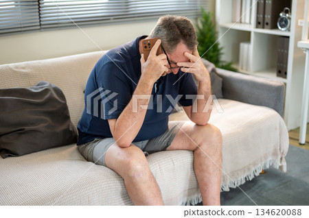 Old white caucasian man touching head in frustration while talking on phone sitting on sofa in house 134620088