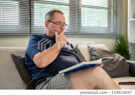 Old white caucasian man wearing glasses looking curious as reading a book sitting on sofa in house. 134620097