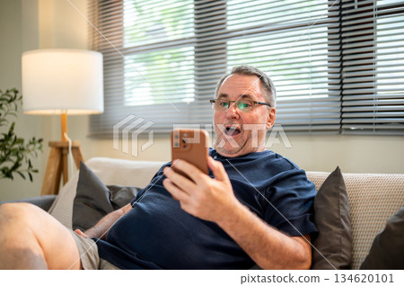 Old white caucasian man wearing glasses looking excited at phone in hand as sitting on sofa in house Old white caucasian man wearing glasses looking excited at phone in hand as sitting on sofa in house 134620101
