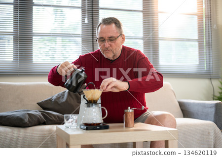 Old white man wearing glasses using kettle pouring hot water into coffee in filter paper on table. 134620119
