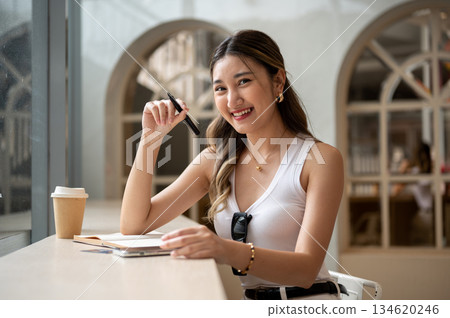 Asian woman in tank top holding pen over notebook with phone and coffee aside sits at wooden table. 134620246