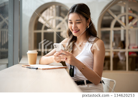 Asian woman smiling while looking at phone holds in hands with notebook and coffee on wooden table. Asian woman smiling while looking at phone holds in hands with notebook and coffee on wooden table. 134620253