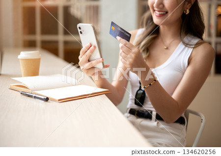 Close up of asian woman holds phone looking at credit card with notebook and coffee on wooden table. Close up of asian woman holds phone looking at credit card with notebook and coffee on wooden table. 134620256