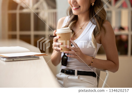 Close up of asian woman in tank top holding coffee in both hands looking at notebook on wooden table Close up of asian woman in tank top holding coffee in both hands looking at notebook on wooden table 134620257