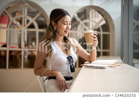 Asian woman in tank top holding coffee cup looking at notebook sits with phone aside on wooden table Asian woman in tank top holding coffee cup looking at notebook sits with phone aside on wooden table 134620258
