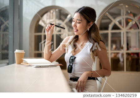 Asian woman in tank top holding a pen looking at notebook sits with phone and coffee on wooden table. Asian woman in tank top holding a pen looking at notebook sits with phone and coffee on wooden table. 134620259
