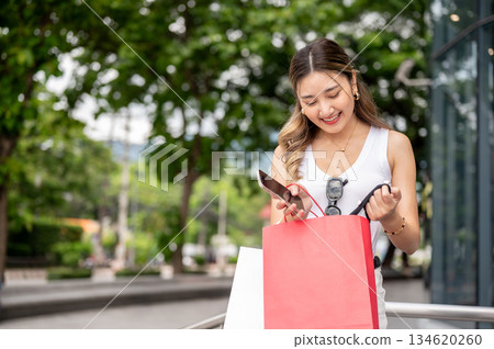 Asian woman in tank top holding looking inside a red shopping bag while standing at mall's forecourt Asian woman in tank top holding looking inside a red shopping bag while standing at mall's forecourt 134620260