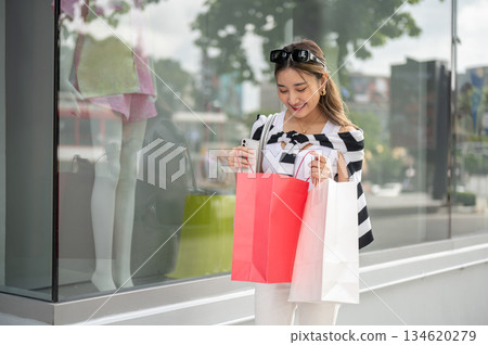 Asian woman looking inside shopping bag standing near a mannequin in mall's glass display cabinet. Asian woman looking inside shopping bag standing near a mannequin in mall's glass display cabinet. 134620279
