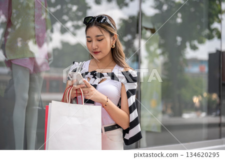 Asian woman holding shopping bag texting on phone near mannequin in the mall's glass display cabinet Asian woman holding shopping bag texting on phone near mannequin in the mall's glass display cabinet 134620295