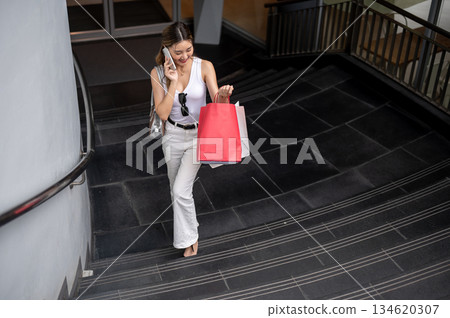 Asian woman in tank top looks at shopping bags during phone call as walking up from mall underground Asian woman in tank top looks at shopping bags during phone call as walking up from mall underground 134620307