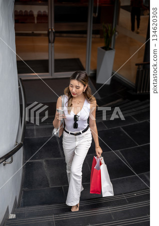 Asian woman in tank top holds shopping bags looking at phone walking upstair from mall underground. 134620308