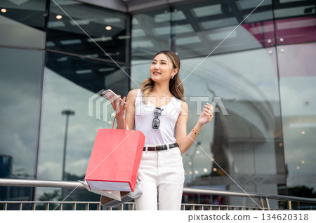 Asian woman in white tank top holding red shopping bag and phone as standing waiting outside a mall. 134620318