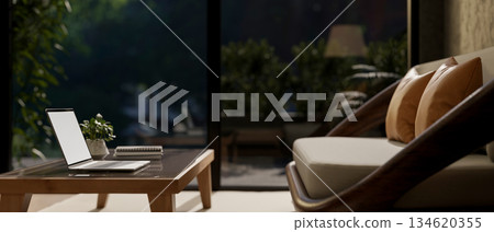 White screen laptop on table in front of a sofa or armchair in living room with glass door at night. 134620355