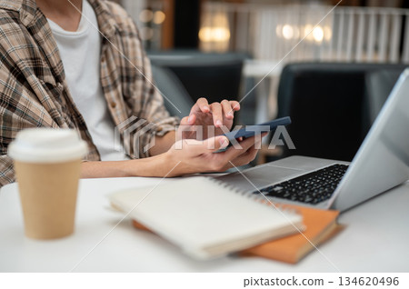 Close up of a man hand holding touching on phone over laptop with coffee and book sits at cafe table 134620496