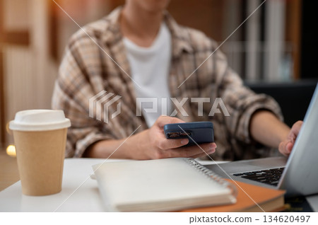 Close up of a man hand holding phone typing on laptop with coffee and notebook sits at cafe table. 134620497
