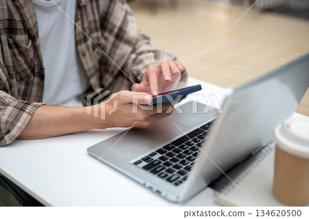 Close up of a man looking touching on phone over laptop with coffee atop book sitting at cafe table. 134620500