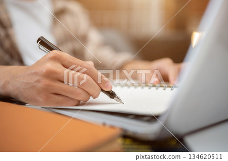 Close up of a man hand holding fountain pen writing in notebook over laptop sitting at table in cafe Close up of a man hand holding fountain pen writing in notebook over laptop sitting at table in cafe 134620511