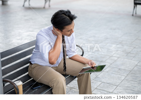 Male office worker massaging or rubbing his neck while holding tablet sitting on steel bench in park 134620587