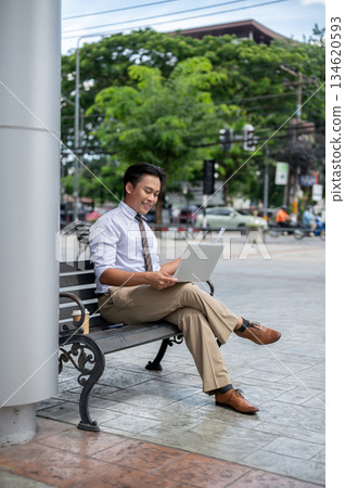 Smiling male office worker looking at his laptop while sitting cross legged on steel bench in park. 134620593