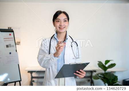 Asian female doctor with stethoscope standing holding clipboard and pointing a pen in diagnosis room 134620729