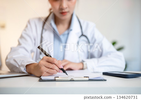 Asian female doctor holding pen writing in paper on clipboard with laptop on table in diagnosis room 134620731