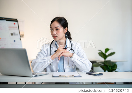 Asian female doctor holding hands and pen under chin looking at laptop on table in diagnosis room. 134620743